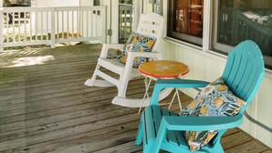 A covered porch with wooden flooring, two white chairs, a turquoise chair, and a small round table. - PRIVATE Pool w/ Lithium  Golf Cart w paid parking INCLUDED. Walk to Restaurants (Oak Island)
