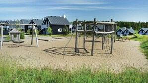 Ein Spielplatz mit Schaukeln und Klettergerüsten, umgeben von Strandhäusern mit blauen Geländern und grauen Dächern.