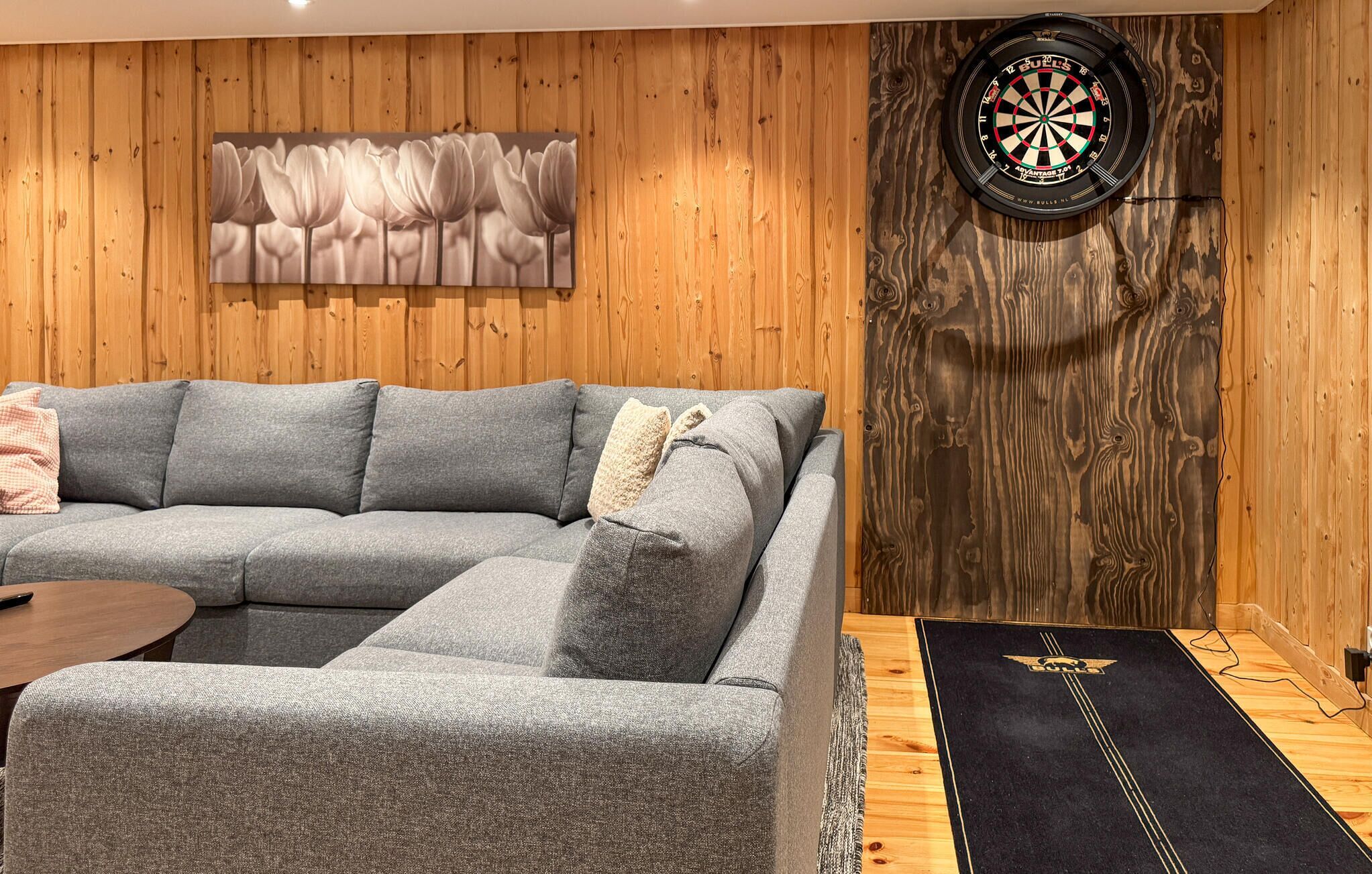 Une pièce avec un canapé d’angle gris, un jeu de fléchettes et un mur en bois orné d’un tableau représentant des motifs floraux.