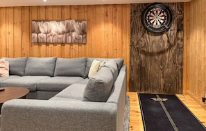 A room with a grey sectional sofa, a dartboard, and a wooden wall with a floral painting. - 5 bedroom awesome home in Kopparberg (Kopparberg)