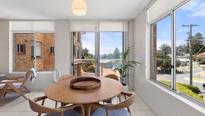 A modern dining area with a round wooden table and chairs, large windows offering a view of a residential area, and a hanging light fixture. - Seaview 6 - Great Views in the Heart of Avoca (Avoca Beach)