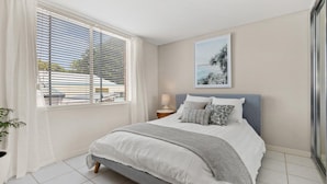 A bedroom with a bed, a nightstand, a lamp, a window with blinds, a potted plant, and a framed picture on the wall. - Seaview 6 - Great Views in the Heart of Avoca (Avoca Beach)