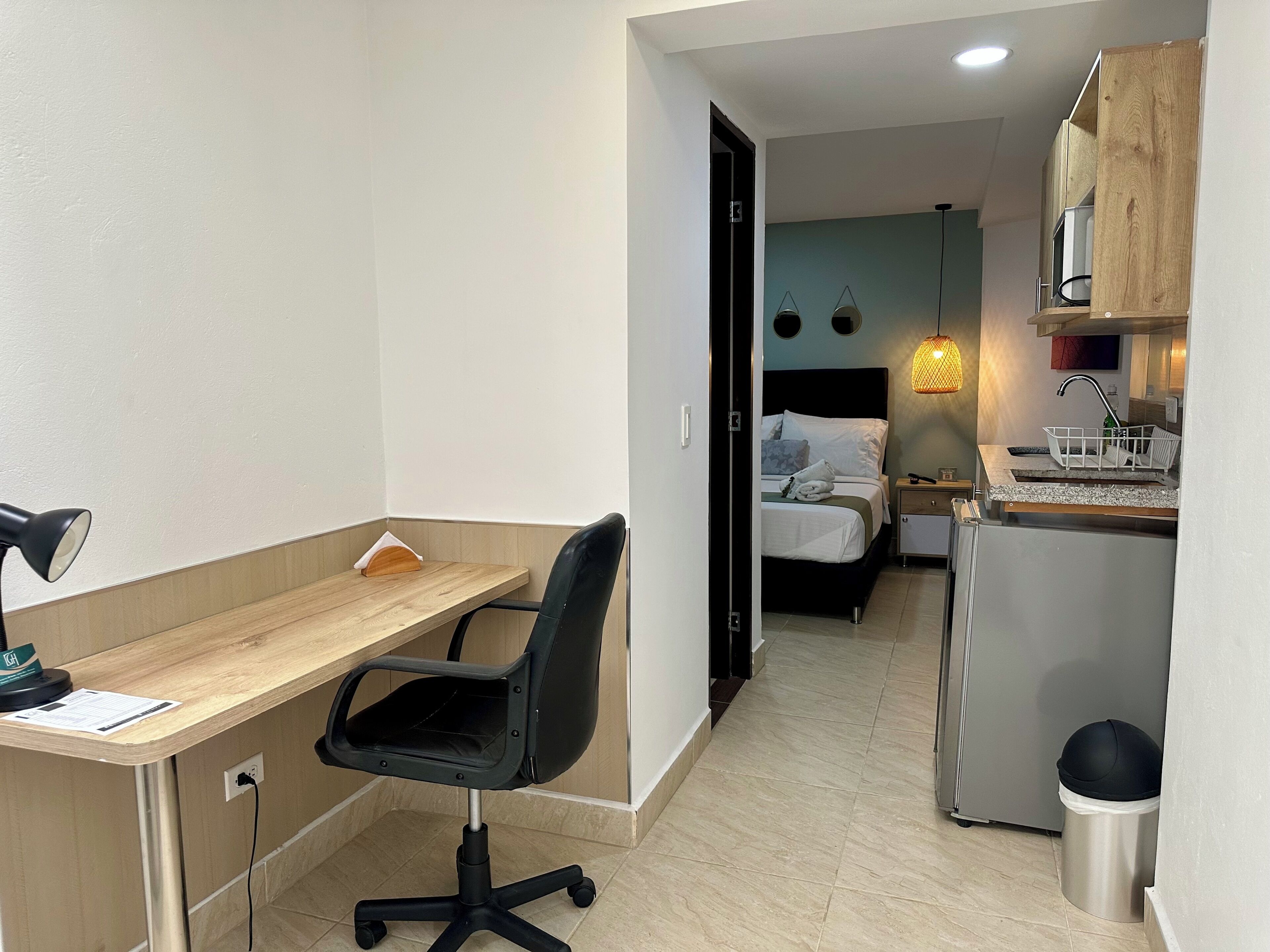 A compact hotel room with a wooden desk, a black office chair, a bed with white bedding, a small kitchenette, and a trash bin.