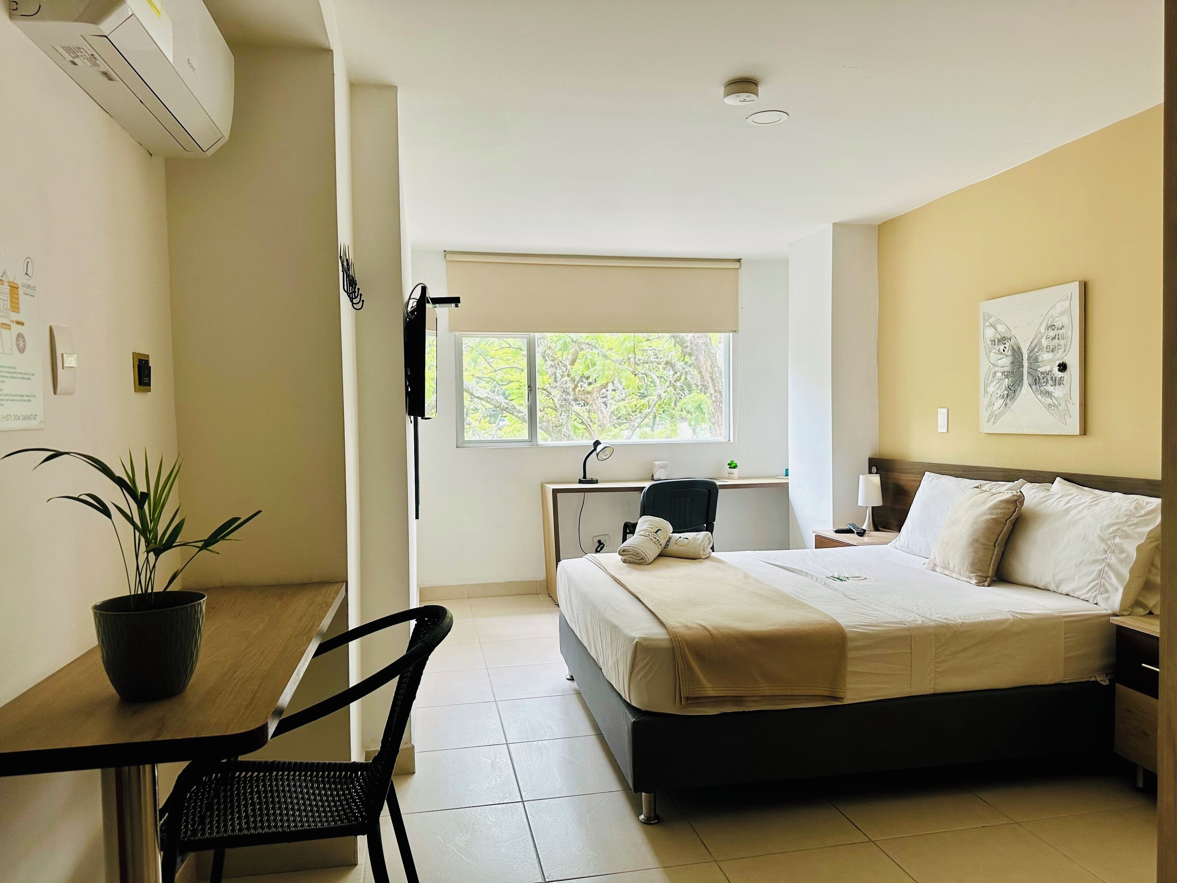 A hotel room with a bed, a desk, a chair, a potted plant, a window, and a ceiling-mounted air conditioner.