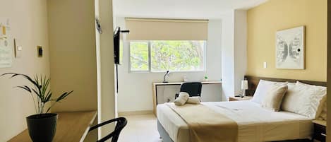 A hotel room with a bed, a desk, a chair, a potted plant, a window, and a ceiling-mounted air conditioner.