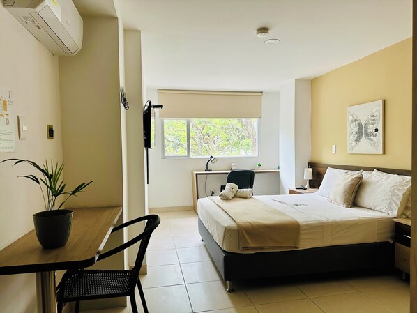 A hotel room with a bed, a desk, a chair, a potted plant, a window, and a ceiling-mounted air conditioner. - Laureles Grand House LGH Hotel - Anteriormente Laureles Guest House (Medellín)