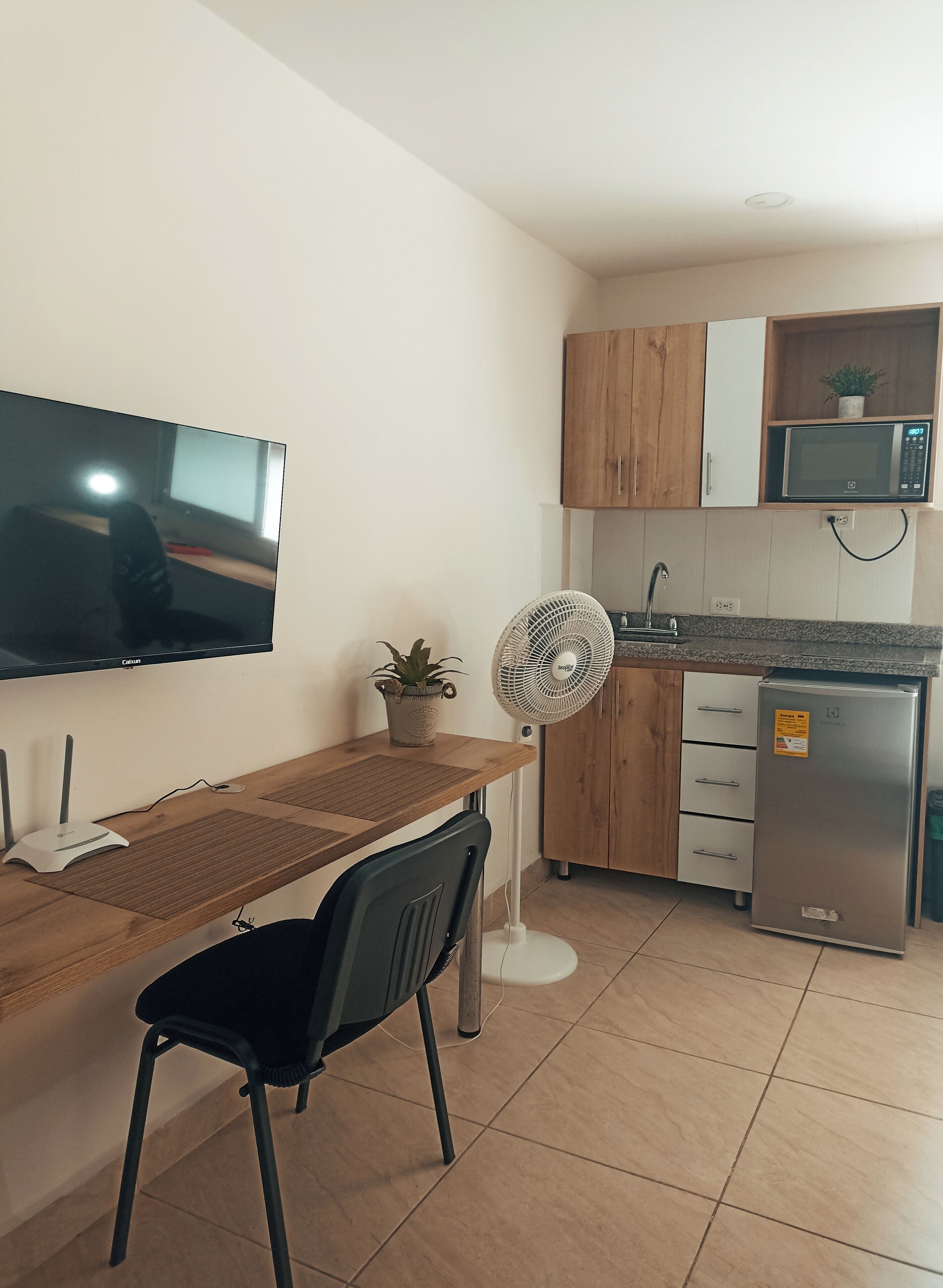 Eine kleine Küche mit Holzausstattung, einer Mikrowelle, einem Kühlschrank, einem Esstisch mit einem Stuhl und einem an der Wand befestigten Fernseher.
