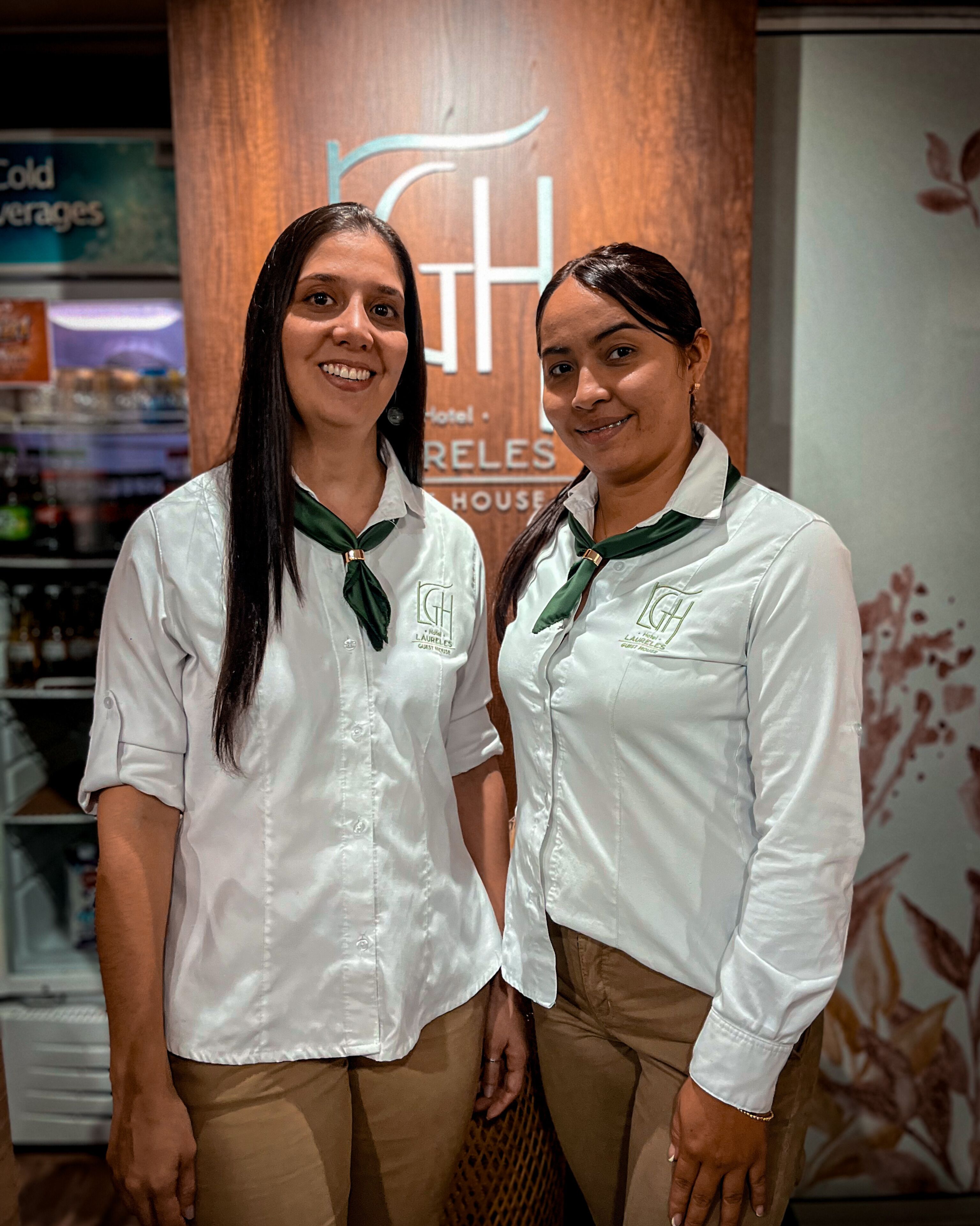 Two individuals in white shirts with green ties standing in front of a wooden wall with a logo and text.