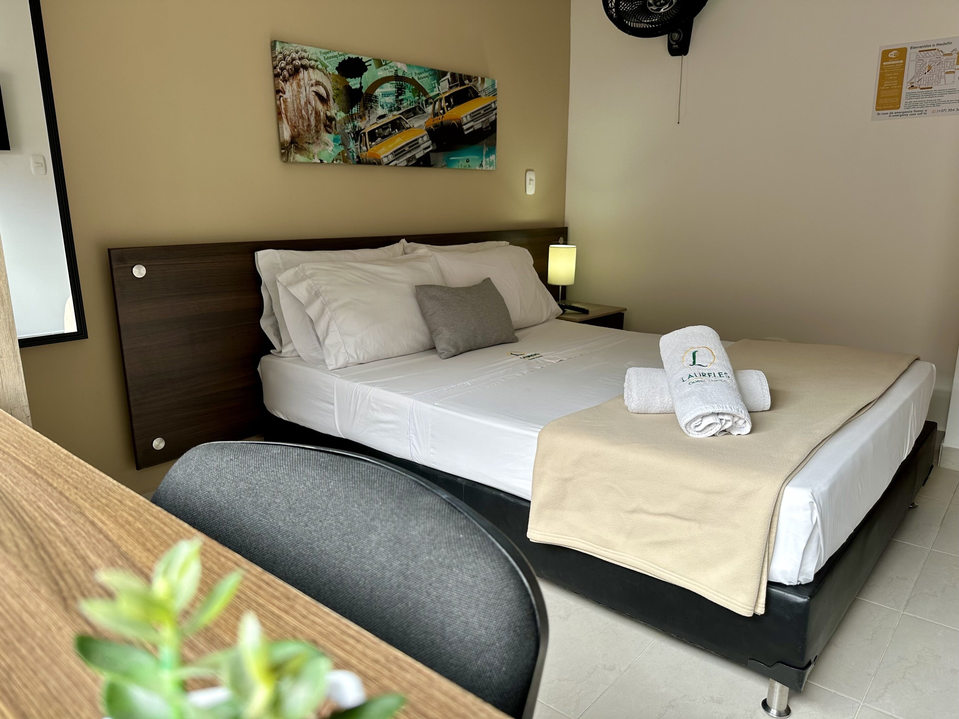 A neatly made bed with a white comforter and pillows, a bedside lamp, a framed artwork on the wall, and a small potted plant on the desk.