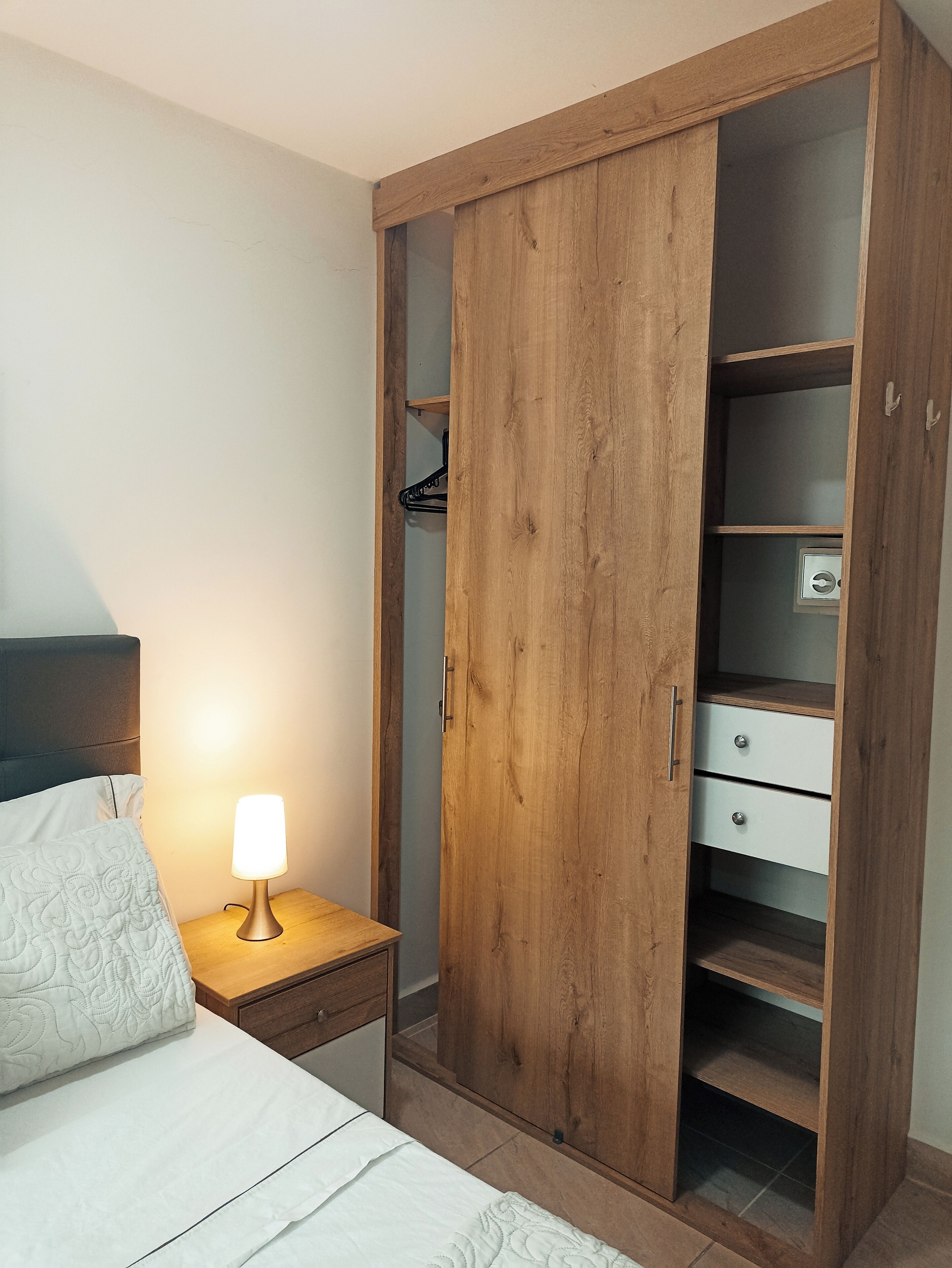 A bedroom with a bed, a nightstand with a lamp, and a wooden wardrobe with open doors.