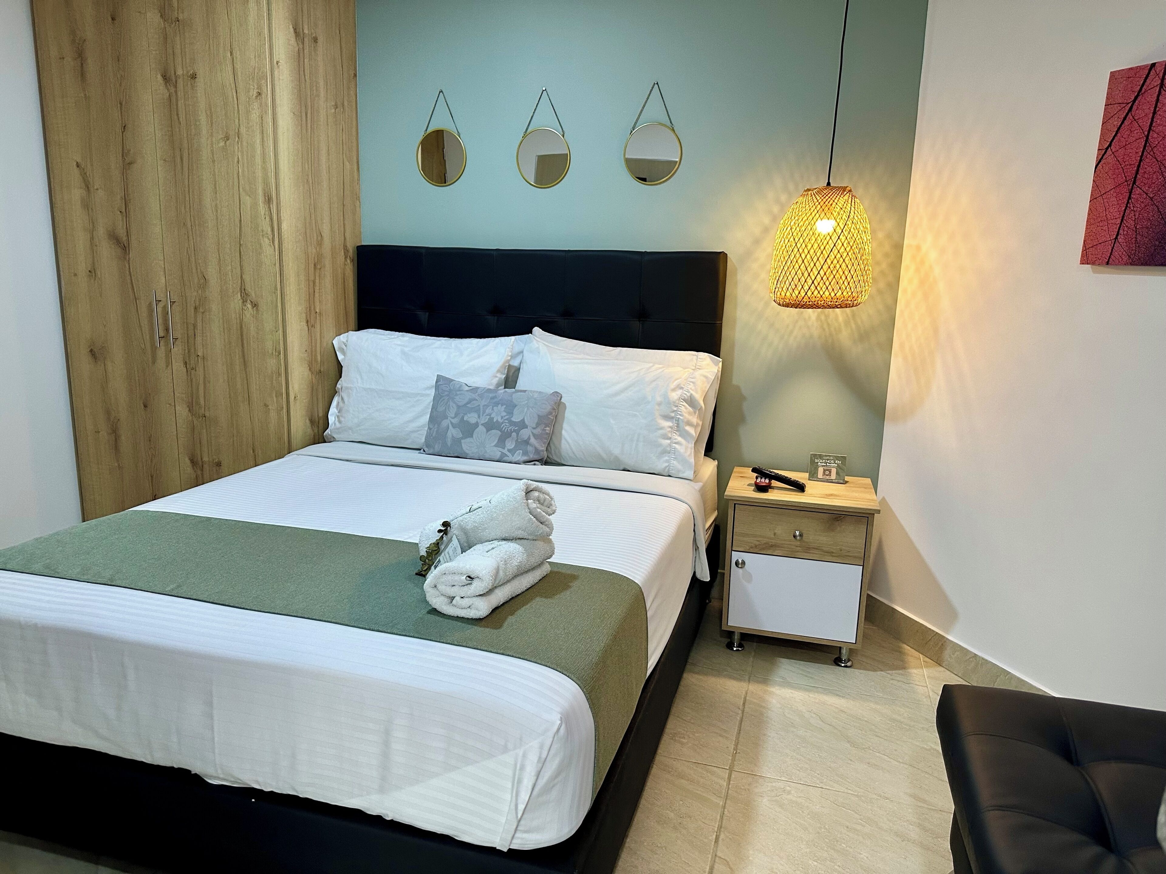 A neatly made bed with a folded towel, a bedside table with a clock, and a hanging light fixture.
