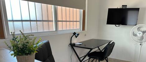 Une petite pièce moderne, dotée d’une table en verre, de chaises en cuir noir, d’une plante en pot, d’une lampe de bureau, d’un téléviseur à écran plat et d’un ventilateur sur pied.