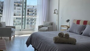 A modern hotel room with a large bed, a seating area with a chair, a small table, and a view of the city through large windows. - Studio β Super (Buenos Aires)