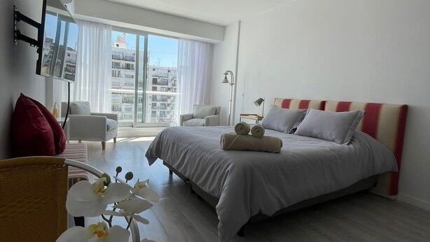 A hotel room with a bed, a chair, a desk, and a view of the city through large windows. - Studio β Super (Buenos Aires)