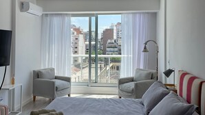 A modern hotel room with a large bed, two armchairs, a TV, and a balcony with city views. - Studio β Super (Buenos Aires)