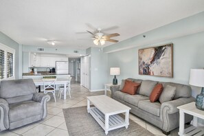 Un vaste espace de vie comprenant un canapé gris, une table basse et une table à manger avec des chaises. On y trouve également un ventilateur de plafond et un tableau accroché au mur.
