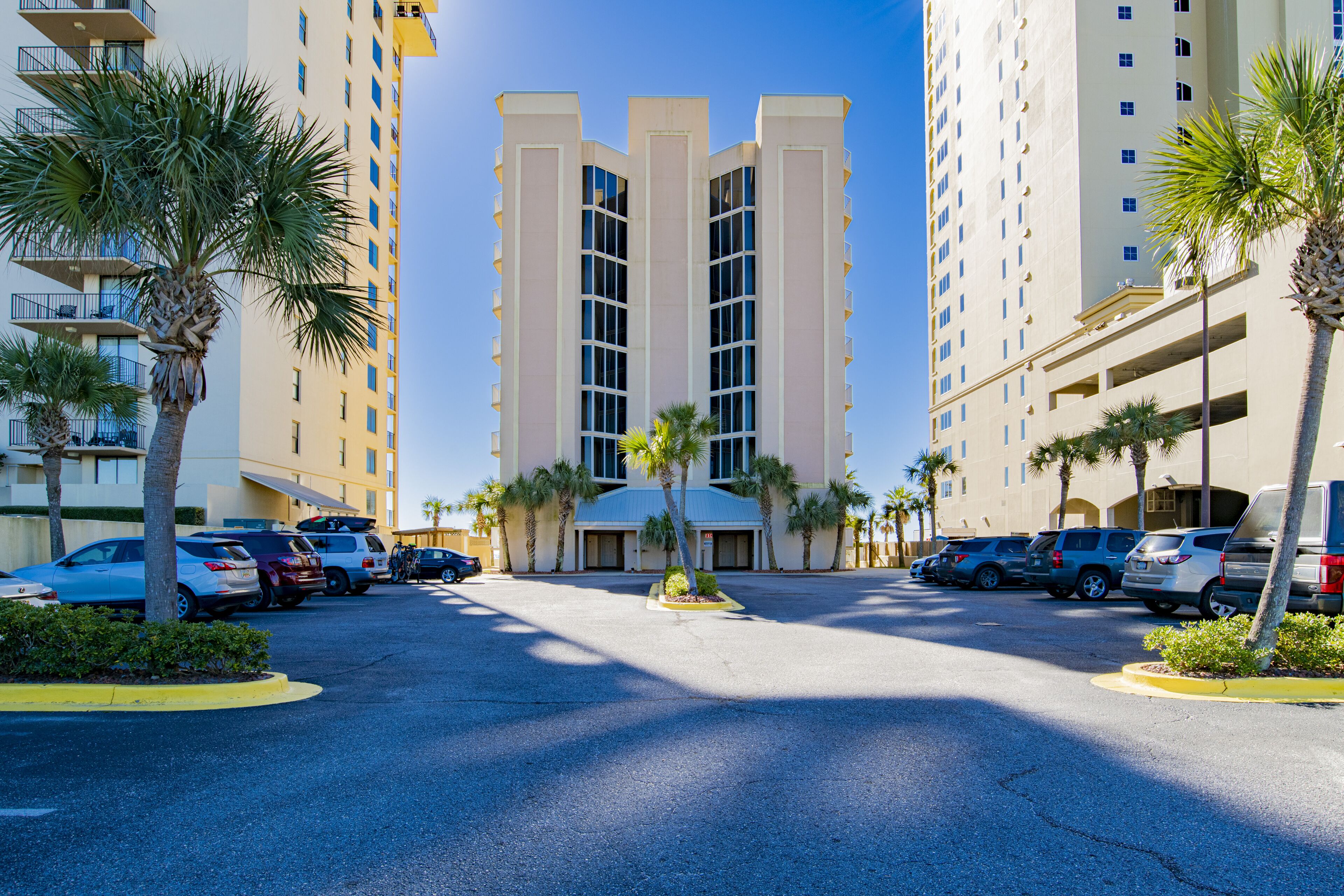 A parking lot with multiple cars, palm trees, and a building with a central tower and balconies.