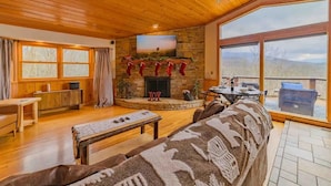A cozy living room with a fireplace, wooden furniture, and large windows offering a view of the outdoors. - Location Location  Location ! Resort Pools , Hot Tub ! MILLION DOLLAR VIEW !! (Gatlinburg)