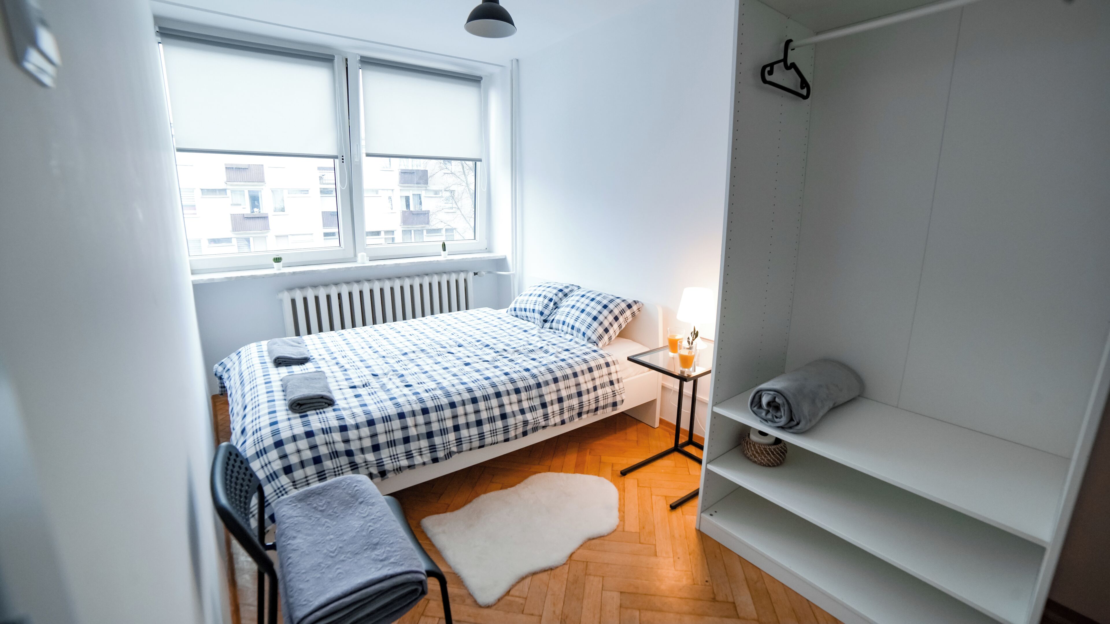 Une petite chambre à coucher, lumineuse, avec un lit, une table de chevet, un radiateur et une fenêtre donnant sur des immeubles.