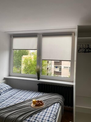 Un dormitorio con cama, ventana con vista a edificios, un radiador y una planta pequeña en el alféizar.