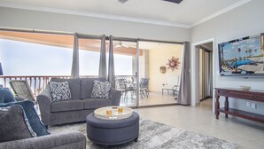 Amplia sala de estar con un sofá gris, un reposapiés y sillones, un comedor con mesa y sillas, un ventilador de techo y un ventanal con vistas al exterior.