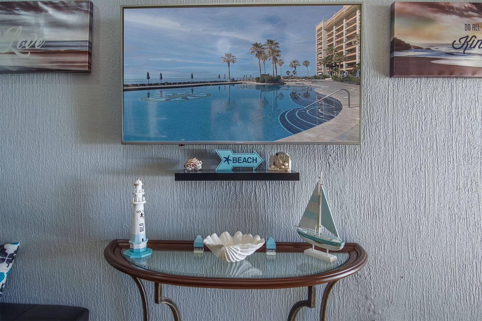 Un téléviseur fixé au mur qui affiche une scène de station balnéaire, une petite étagère avec un panneau décoratif sur le thème de la plage, et une table en verre agrémentée d’objets décoratifs sur le thème de la mer.