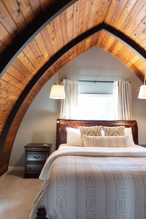 Ein Schlafzimmer mit Holzboden, einem Bett mit gemusterten Textilien, einem Nachttisch und einem Fenster mit Vorhängen.