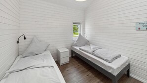 Une petite pièce aux murs recouverts de lambris blancs, avec deux lits simples, une table de chevet, une lampe et une fenêtre.
