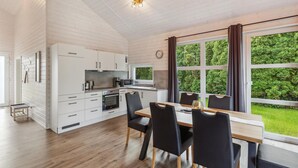 Une cuisine moderne avec des meubles blancs, un coin repas doté d’une table en bois et de chaises noires, et de grandes fenêtres offrant une vue sur la verdure.