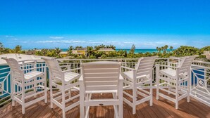 Une terrasse avec des chaises blanches et une piscine, donnant sur une plage bordée de palmiers et surplombant un ciel bleu azur.