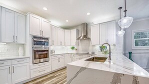 Une cuisine moderne avec des meubles blancs, des appareils en acier inoxydable, un îlot en marbre et des luminaires suspendus.