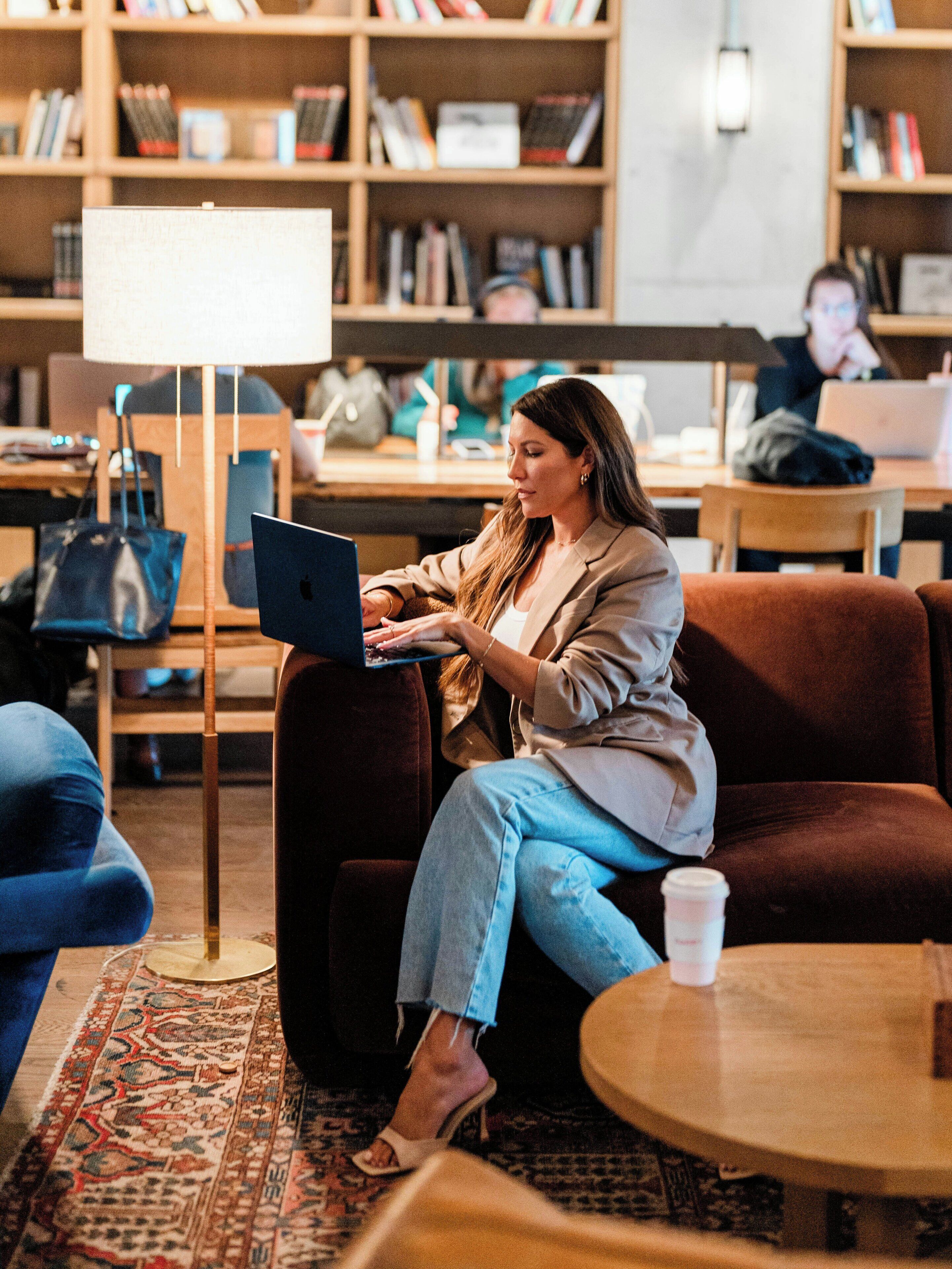 Una persona sentada en un sofá usando una computadora portátil en una habitación con una estantería, una lámpara y una taza de café sobre una mesa.