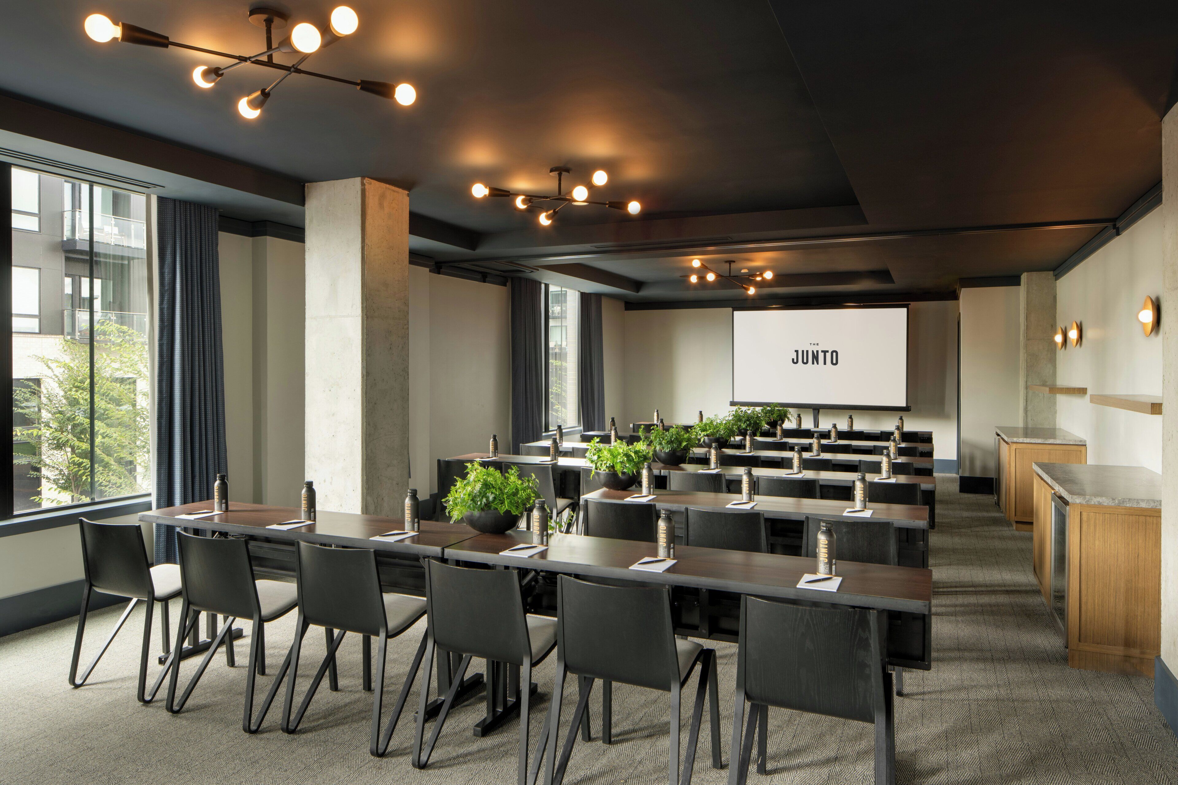 A conference room with multiple tables, chairs, a projector screen, and potted plants.