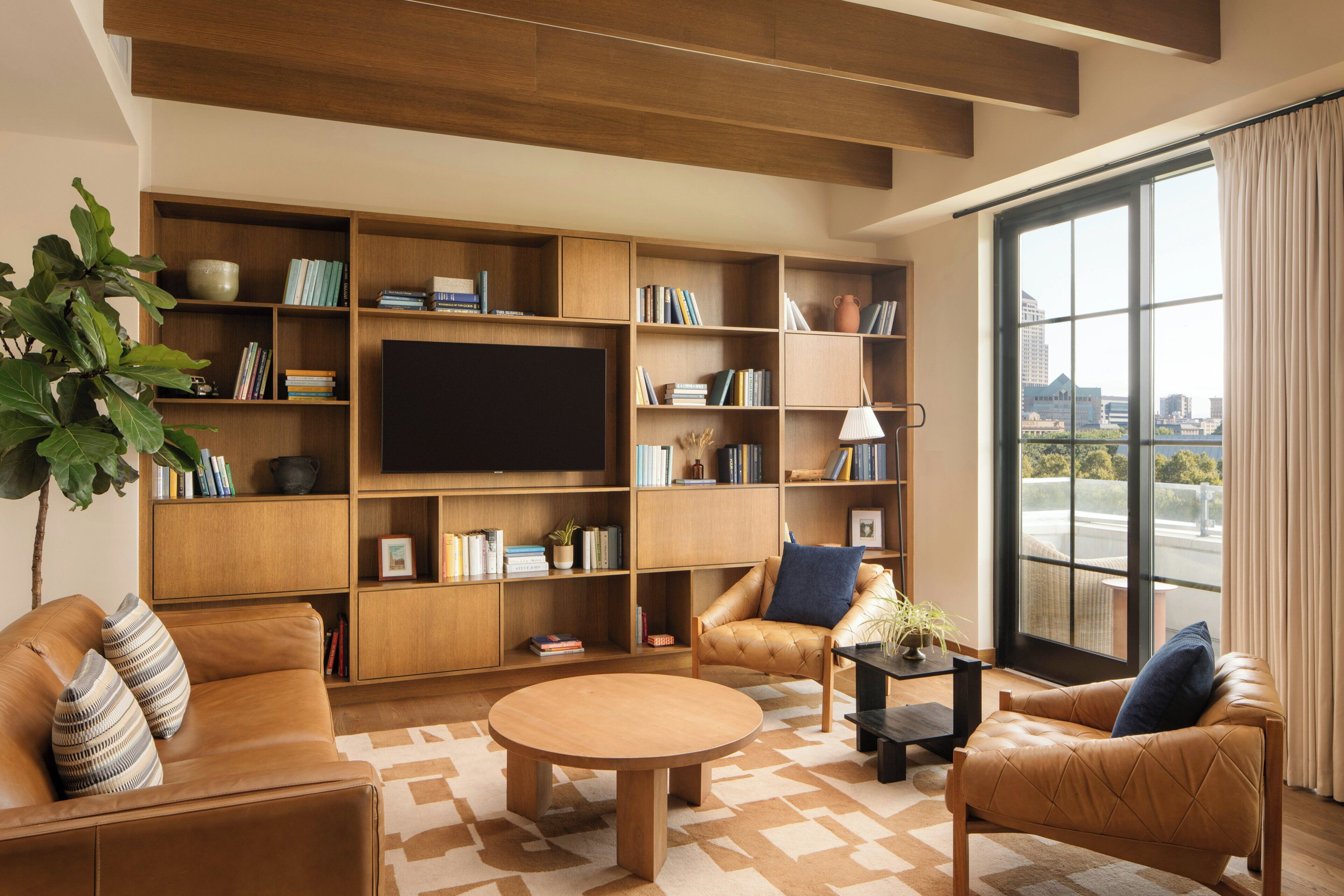 A modern living room with a large TV, wooden bookshelves, leather sofas, and a large window with a city view.