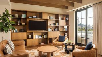 A modern living room with a large TV, wooden bookshelves, leather sofas, and a large window with a city view.