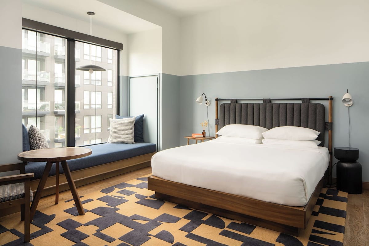A modern hotel room with a large bed, a seating area by the window, and a small table.