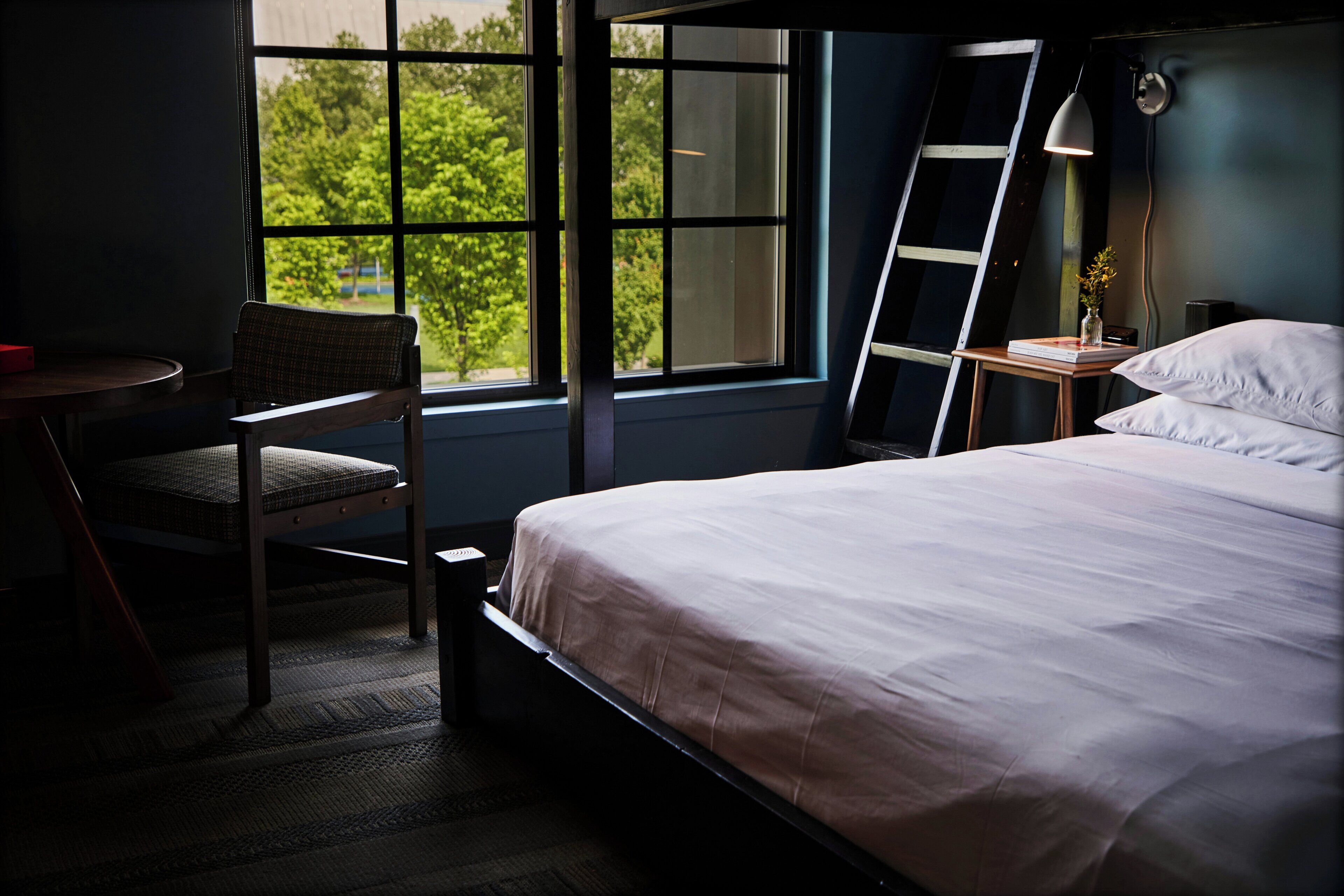 A bedroom with a bed, a chair, a desk, a ladder, and a window with a view of trees.