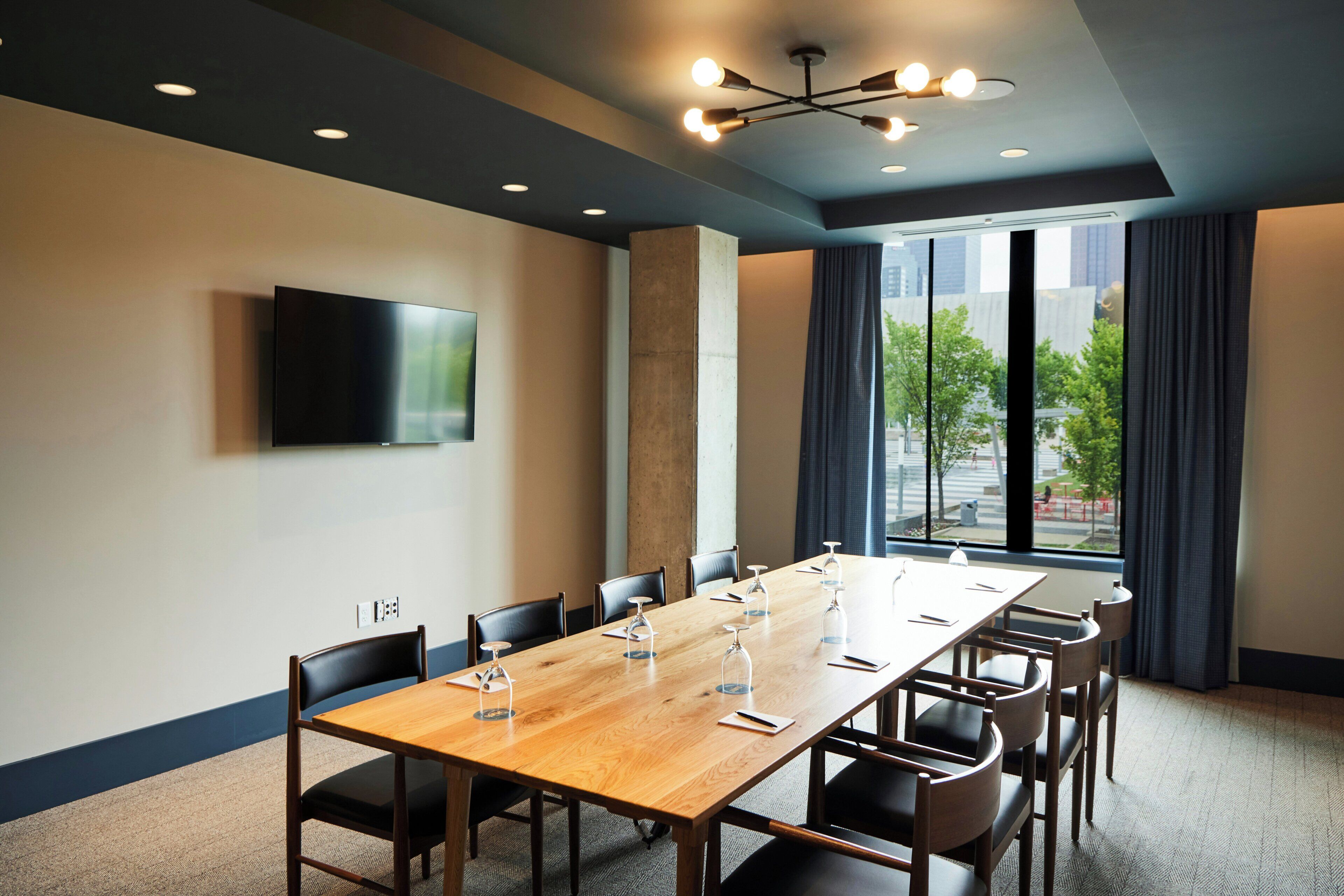 A conference room with a long table, chairs, a flat-screen TV, and a large window with a view of buildings and trees.