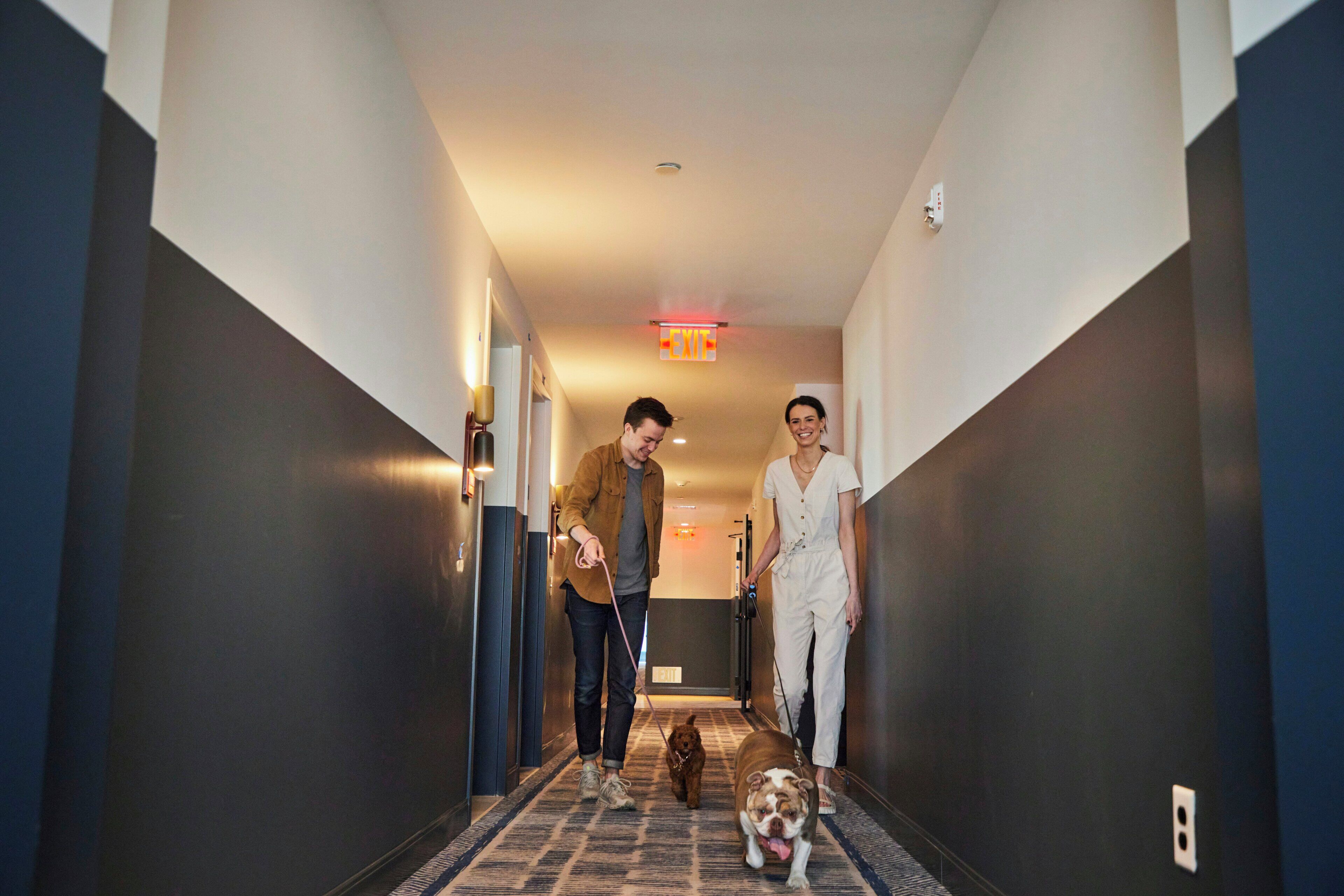 A hotel hallway with a carpeted floor, a wall-mounted exit sign, and a door at the end.