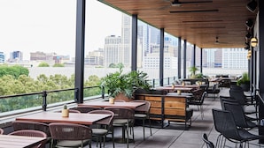 Un espacio exterior para comer con mesas y sillas, ventanas grandes y vistas al horizonte de la ciudad.