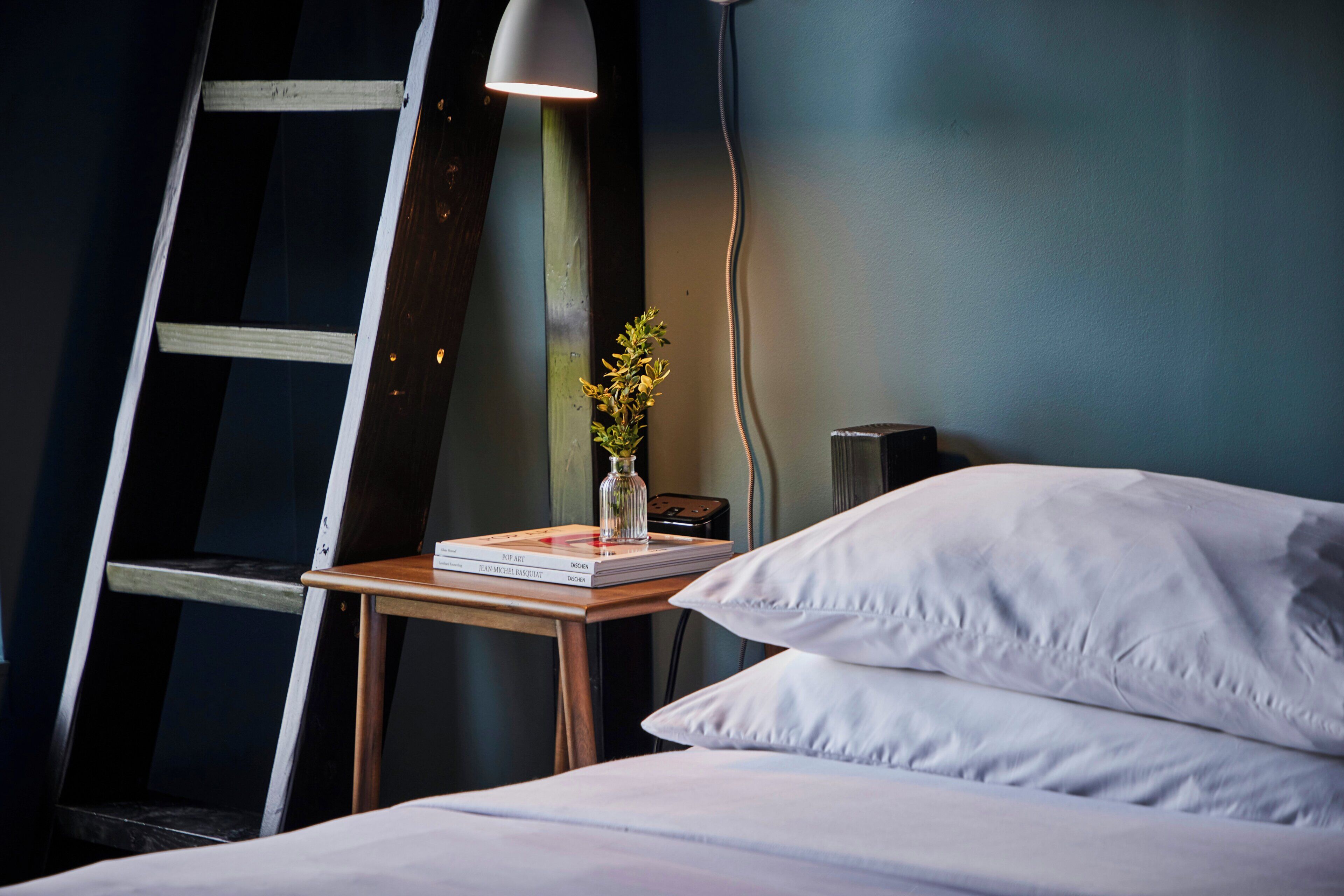 Una cama bien hecha con sábanas blancas, una mesita de noche con una lámpara, un libro y un pequeño jarrón con flores.