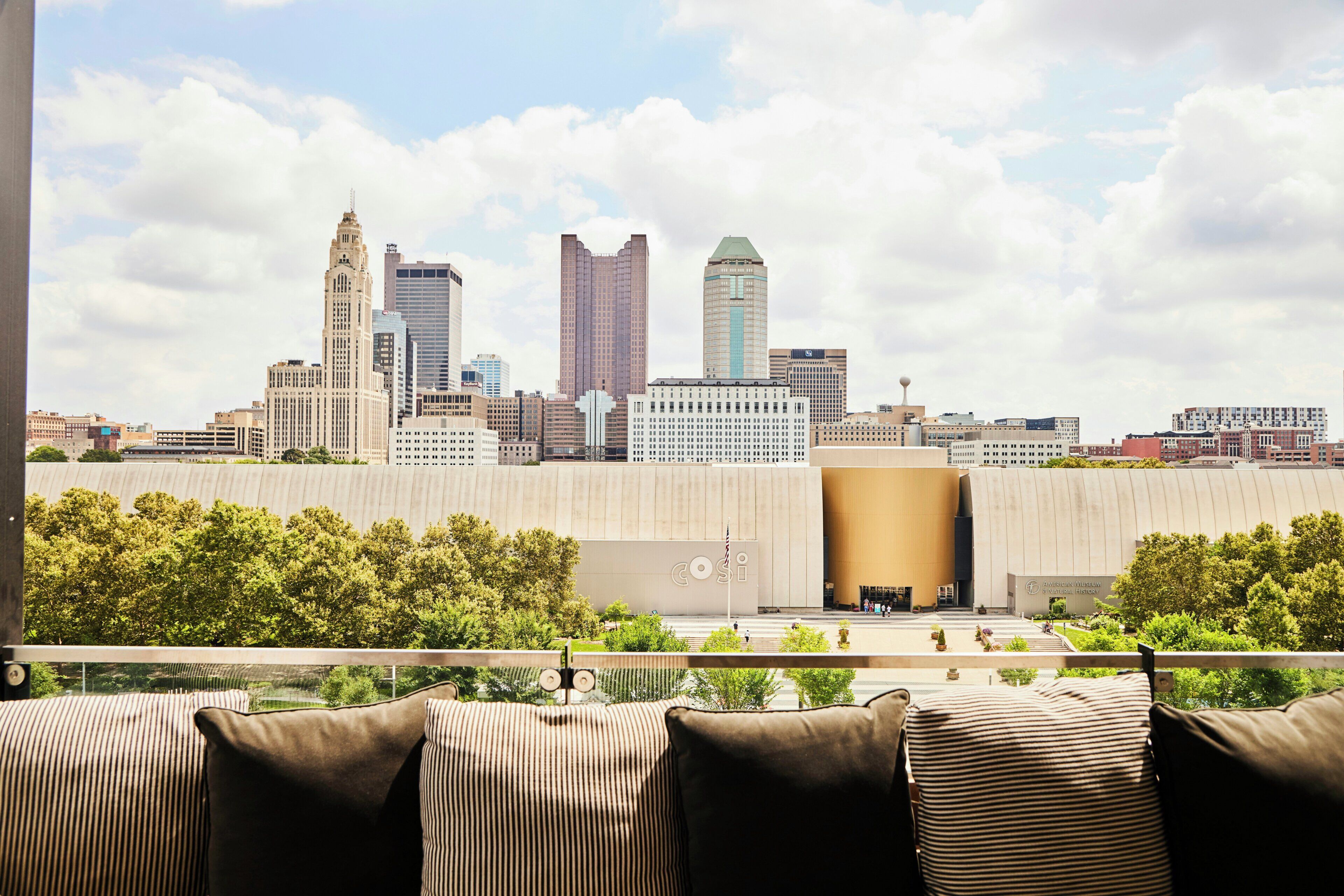 A city skyline with modern buildings, a large open space with a low wall, and a seating area with striped cushions.