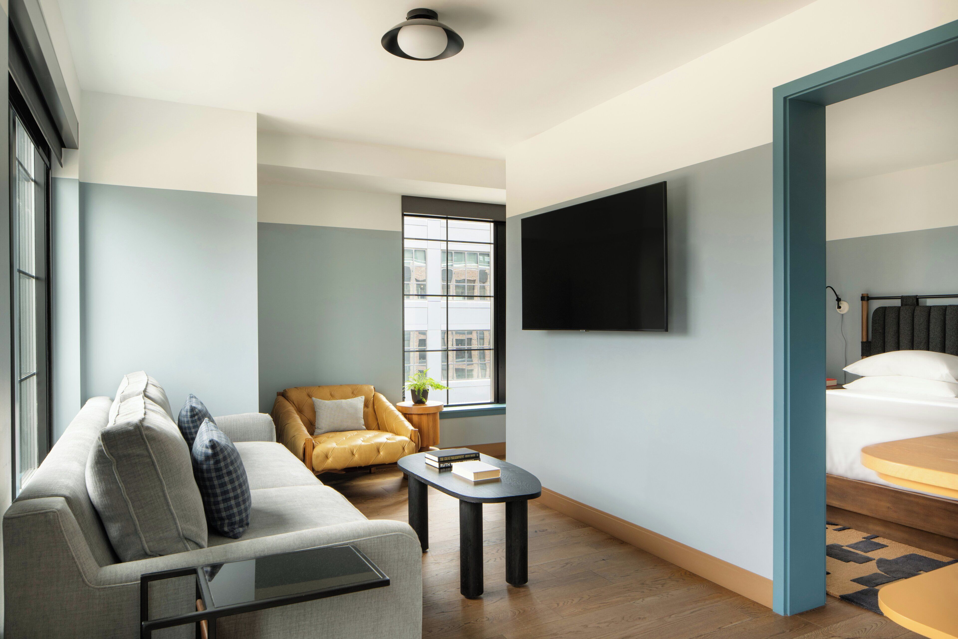 A modern hotel room with a sofa, a small table, a chair, a bed, a TV, and a window with a view of buildings.
