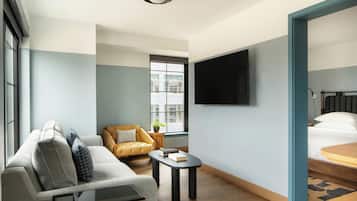 Ein modernes Hotelzimmer mit Sofa, kleinem Tisch, Stuhl, Bett, Fernseher und Fenster mit Blick auf Gebäude.