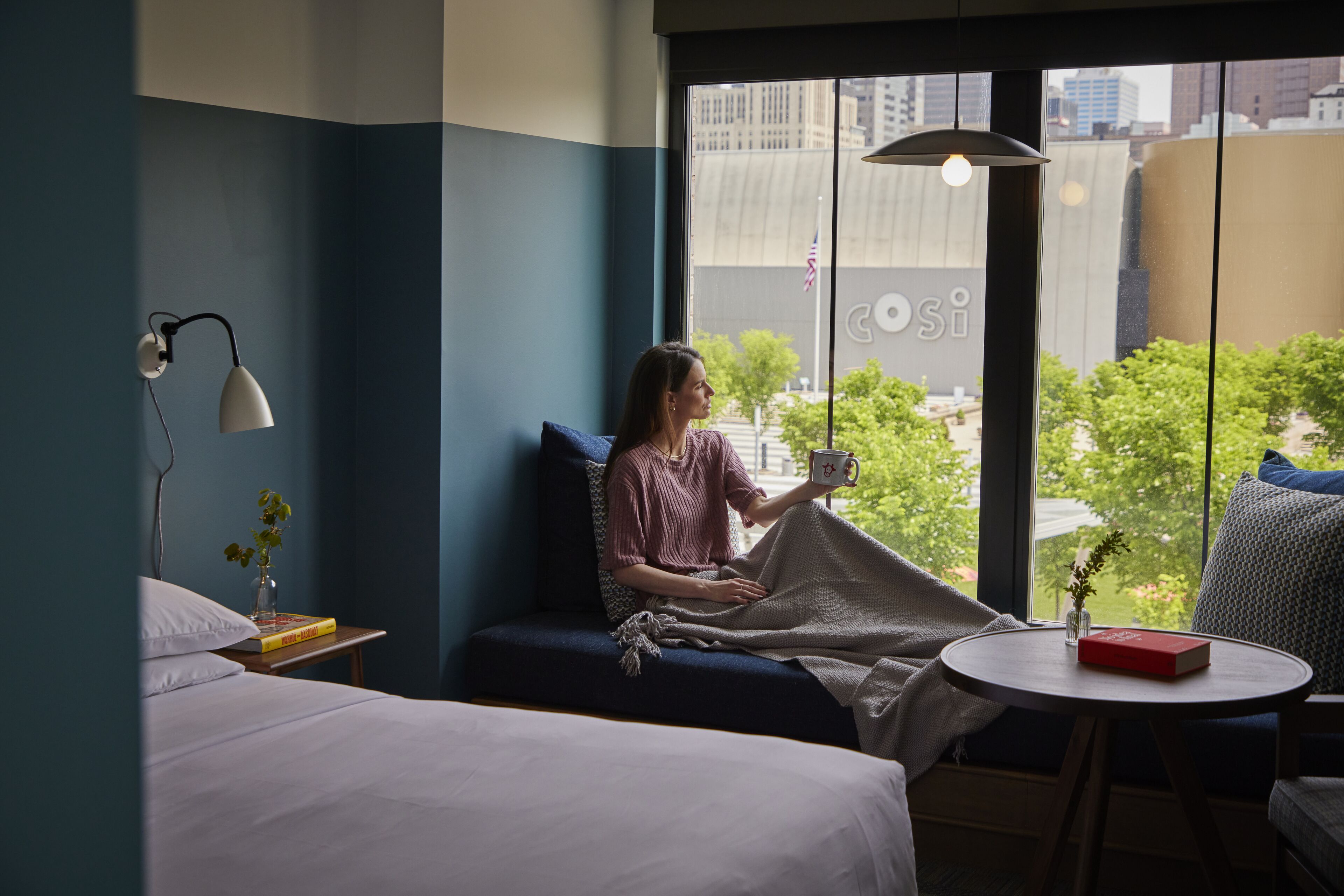 A room with a bed, a sofa, a small table, and a window with a view of buildings and trees.