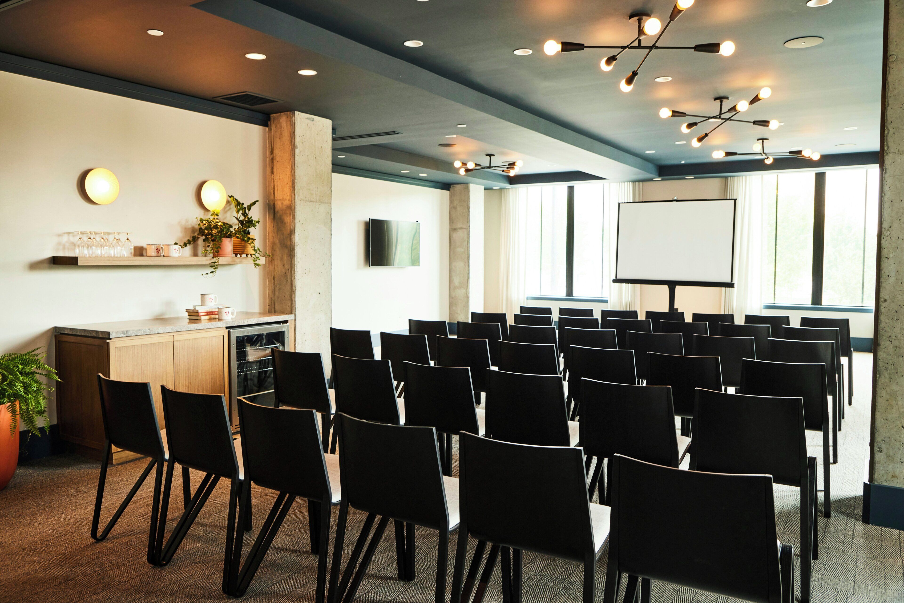 Un salón de conferencias con filas de sillas, una pantalla de proyector y una pequeña área de bar con vasos y una planta.