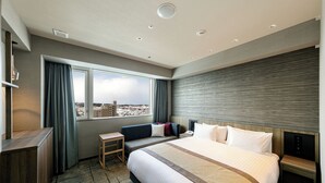 A hotel room with a large bed, a desk, a chair, a nightstand, and a view of the city. - HOTEL VISCHIO TOYAMA by GRANVIA (Toyama)