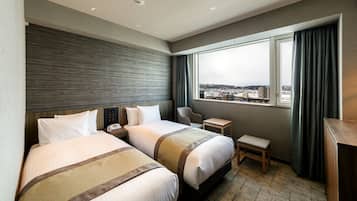 A hotel room with two beds, a small desk, a chair, and a window with a view of a cityscape.