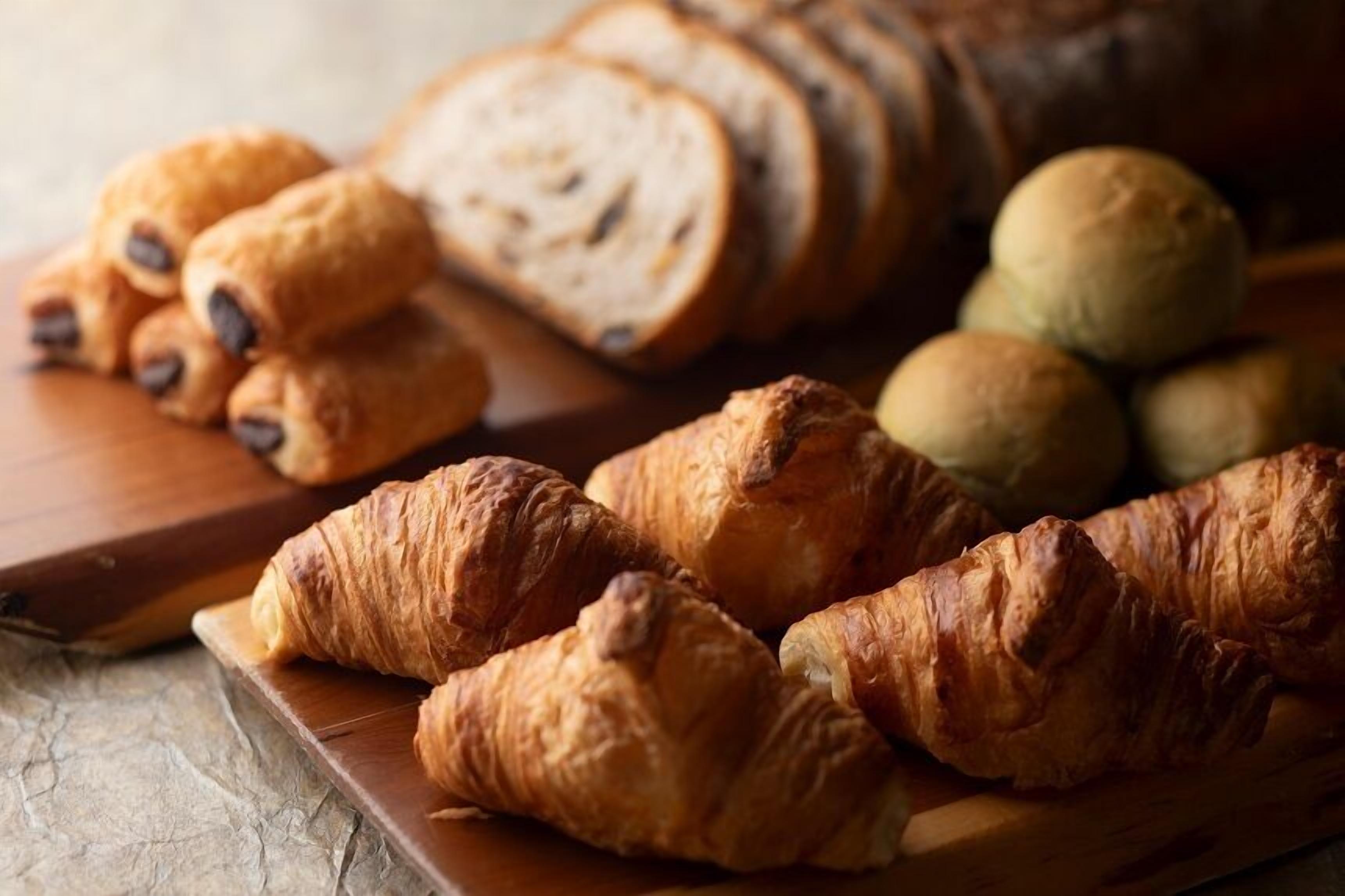 A wooden board with croissants, sliced bread, and round pastries.