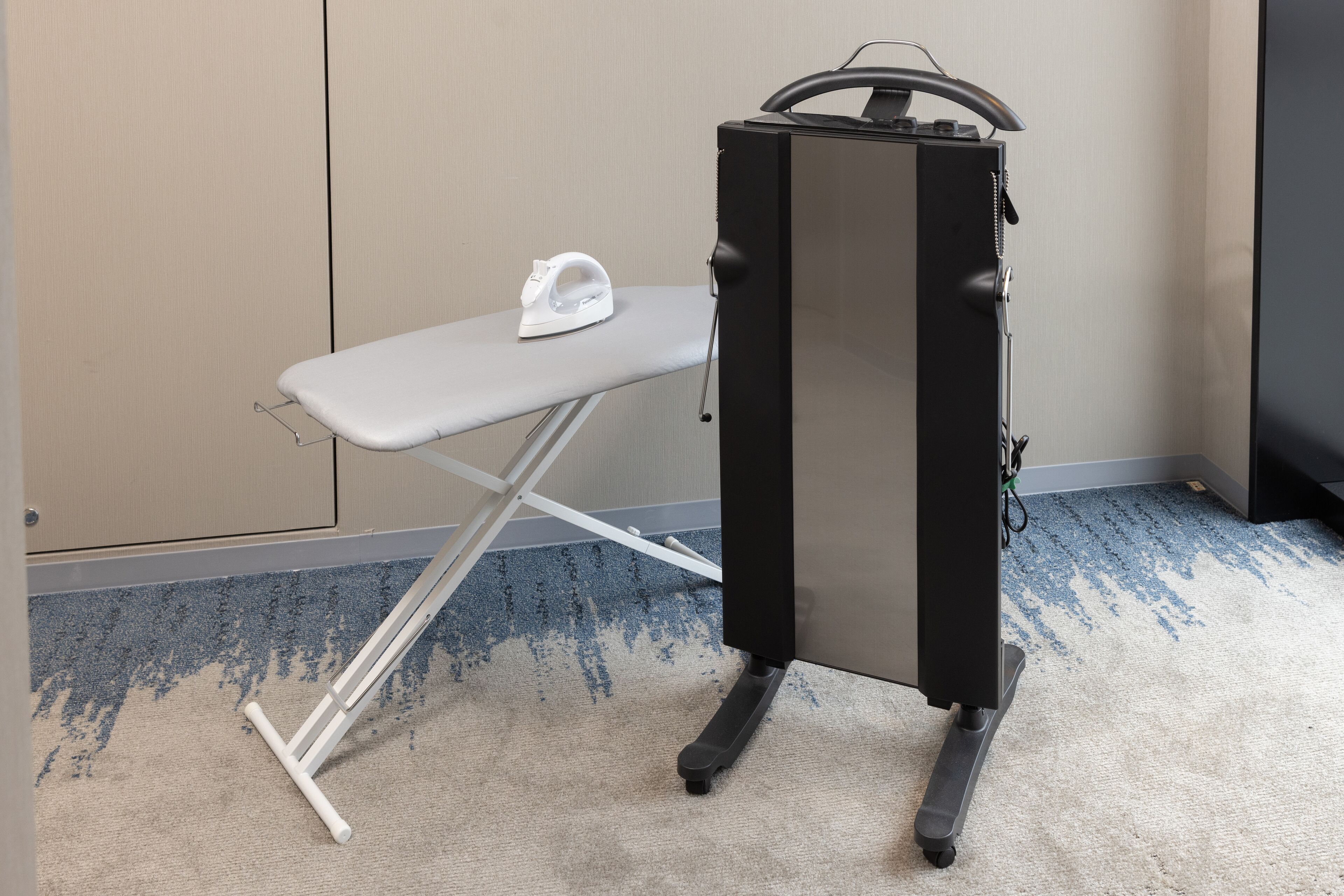 A compact iron and ironing board in a hotel room with a carpeted floor and a plain wall.